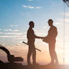 Two construction workers in hard hats shaking hands on a job site with excavator and crane silhouetted against sunset sky
