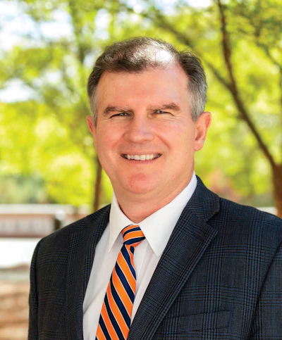 Professional headshot of a smiling man in dark blazer and orange striped tie against green foliage background