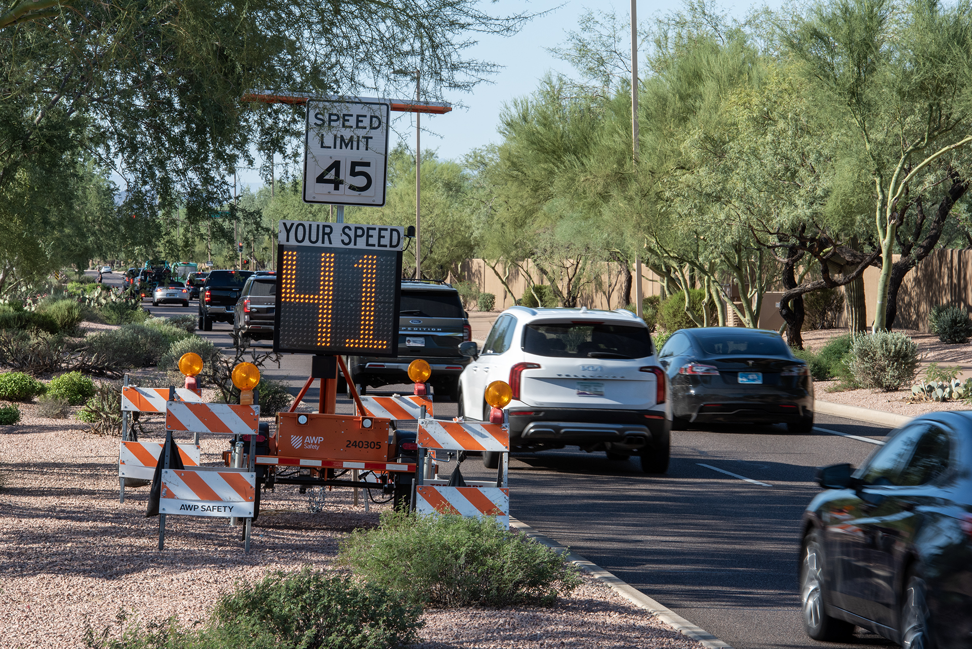 Speed monitoring sign displaying vehicle speeds on a residential street with construction barriers and parked cars