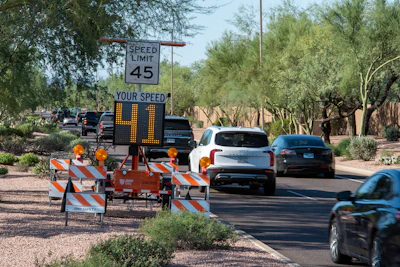 Speed monitoring sign displaying vehicle speeds on a residential street with construction barriers and parked cars