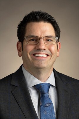 Professional headshot of a smiling man wearing glasses, dark blazer, and blue tie