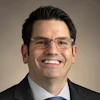 Professional headshot of a smiling man wearing glasses, dark blazer, and blue tie