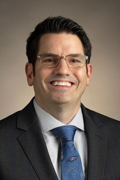 Professional headshot of a smiling man wearing glasses, dark blazer, and blue tie