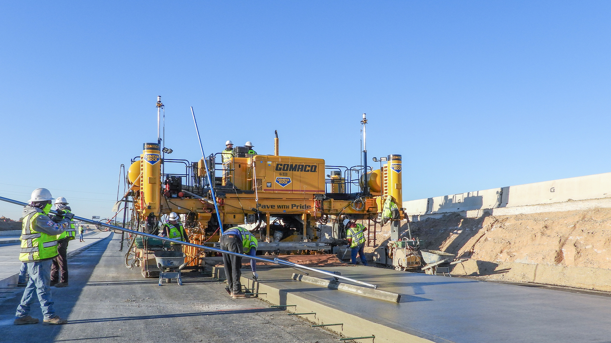 Construction workers operating yellow Gomaco concrete paving machine on highway project with clear blue sky