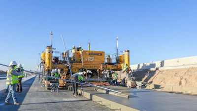 Construction workers operating yellow Gomaco concrete paving machine on highway project with clear blue sky