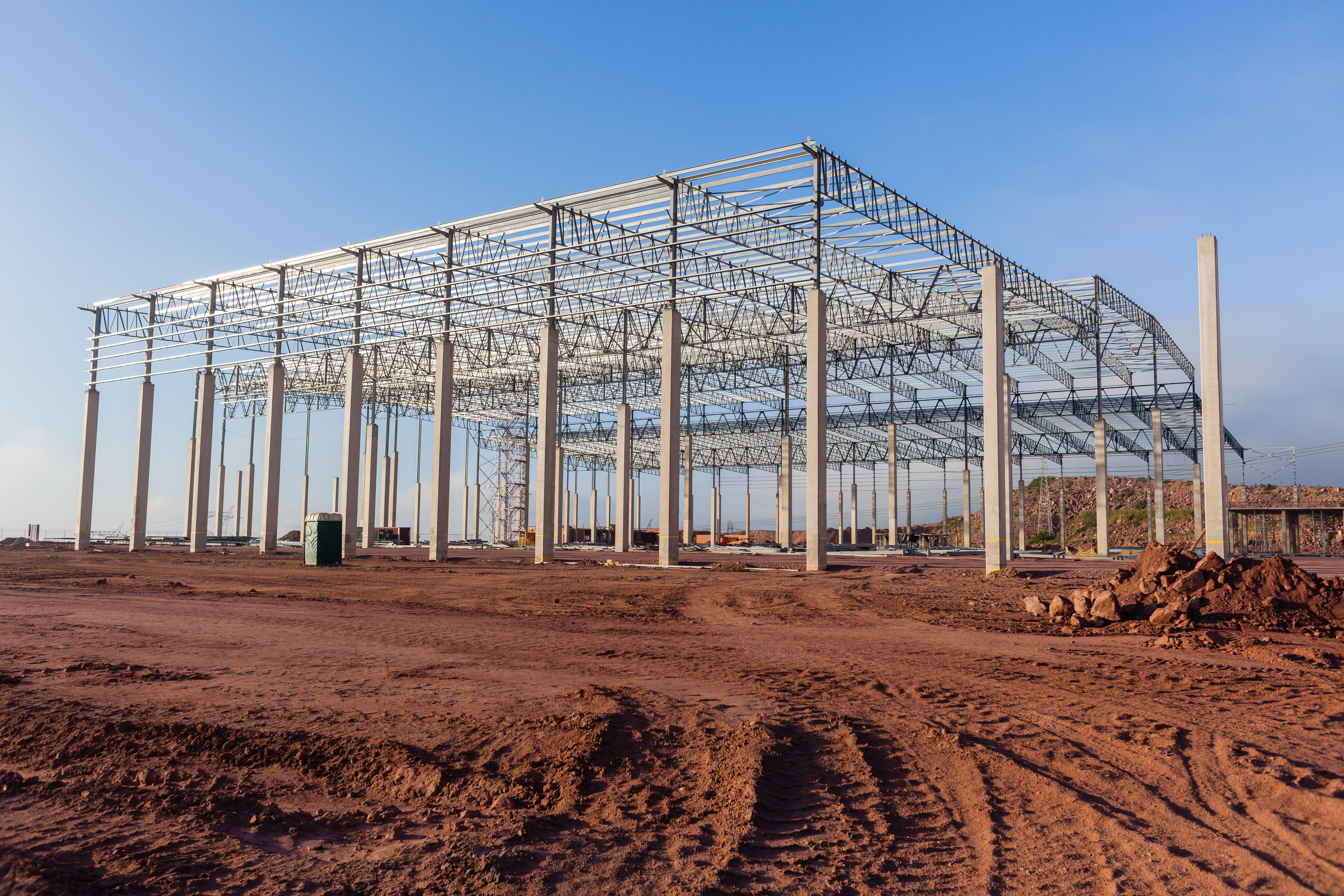Large steel-frame greenhouse structure under construction on bare earth with clear blue sky