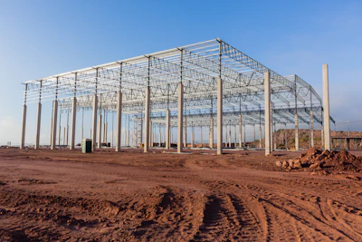 Large steel-frame greenhouse structure under construction on bare earth with clear blue sky
