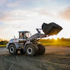 Blue and white wheel loader with raised bucket on rural road at sunset with green fields in background