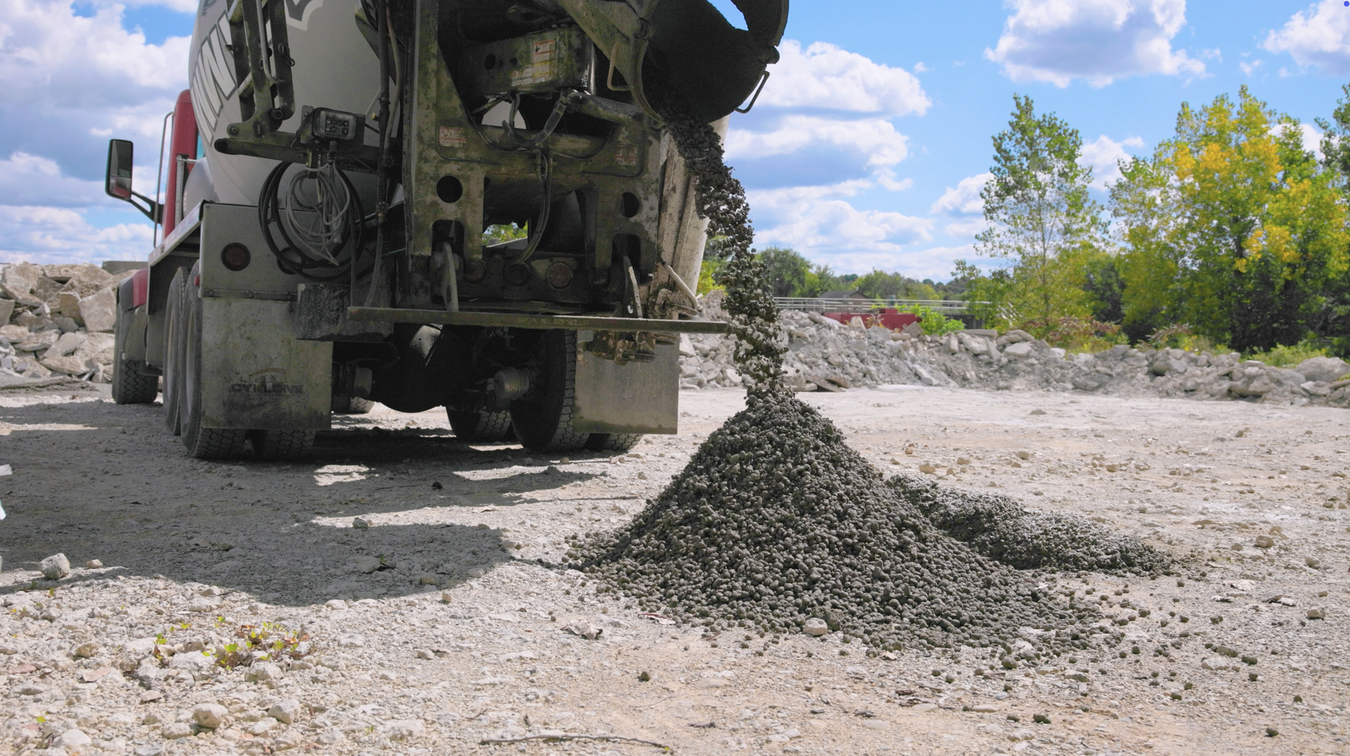 Concrete mixer truck discharging aggregate at construction site with stockpiles and trees