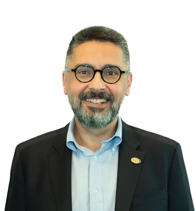 Professional headshot of a smiling man wearing glasses, light blue shirt, and black blazer with a yellow pin