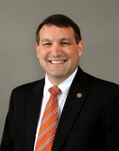 Professional headshot of a smiling man in black suit, white shirt, and orange striped tie against gray background
