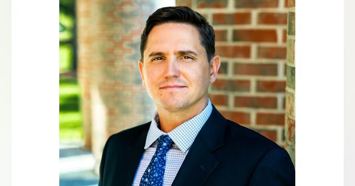 Professional headshot of a man in dark blazer and light blue checkered shirt against brick background Headshot
