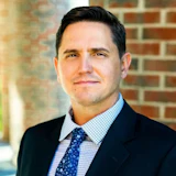 Professional headshot of a man in dark blazer and light blue checkered shirt against brick background
