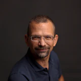 Professional headshot of a man wearing glasses and dark polo shirt against neutral background