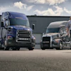 Two Mack trucks parked at an industrial facility with a worker standing between them under a cloudy sky