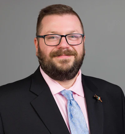Professional headshot of bearded man wearing glasses, pink shirt, and blue tie with black blazer