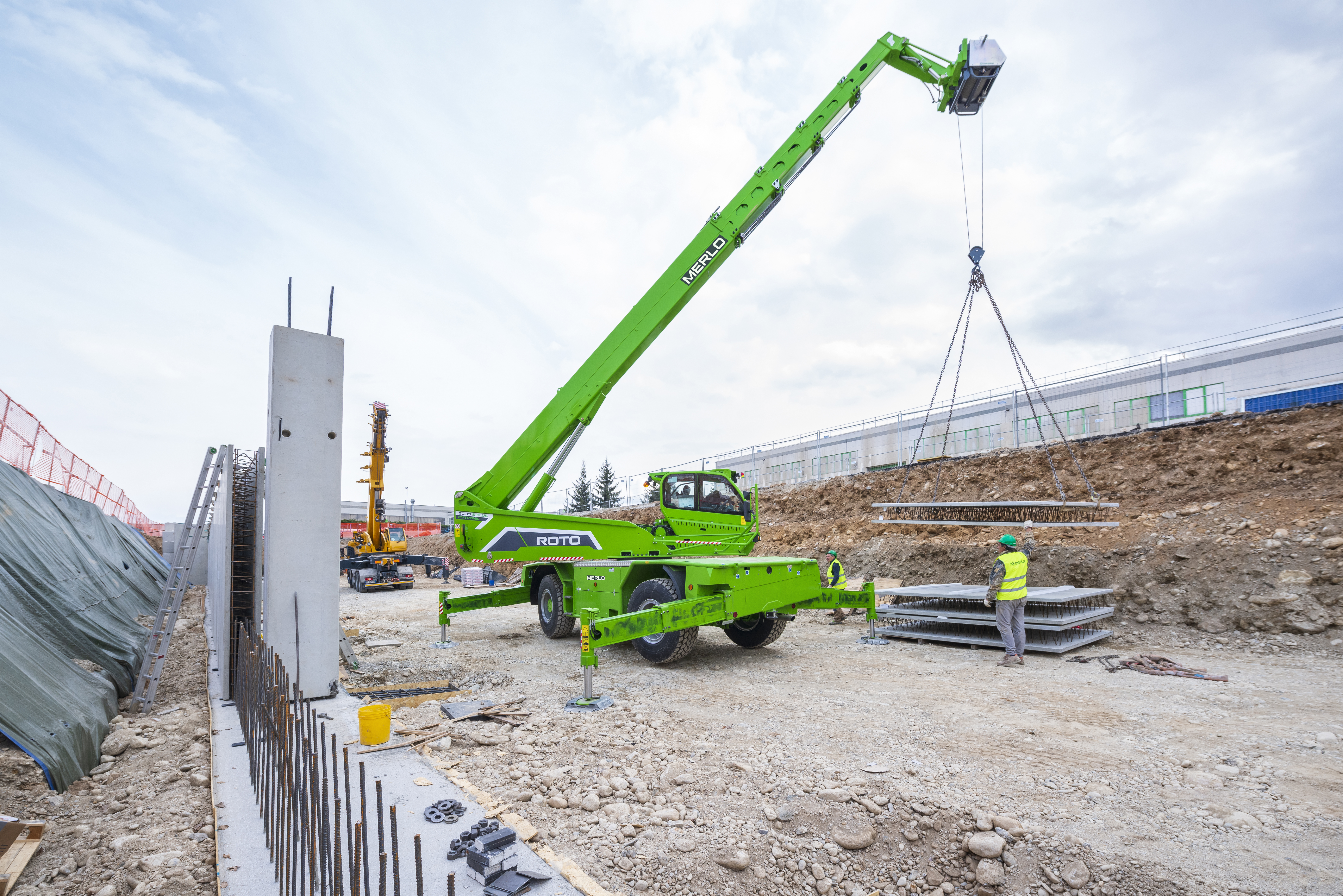 Green MERLO telescopic handler lifting construction materials at a building site with workers and equipment visible