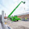Green MERLO telescopic handler lifting construction materials at a building site with workers and equipment visible