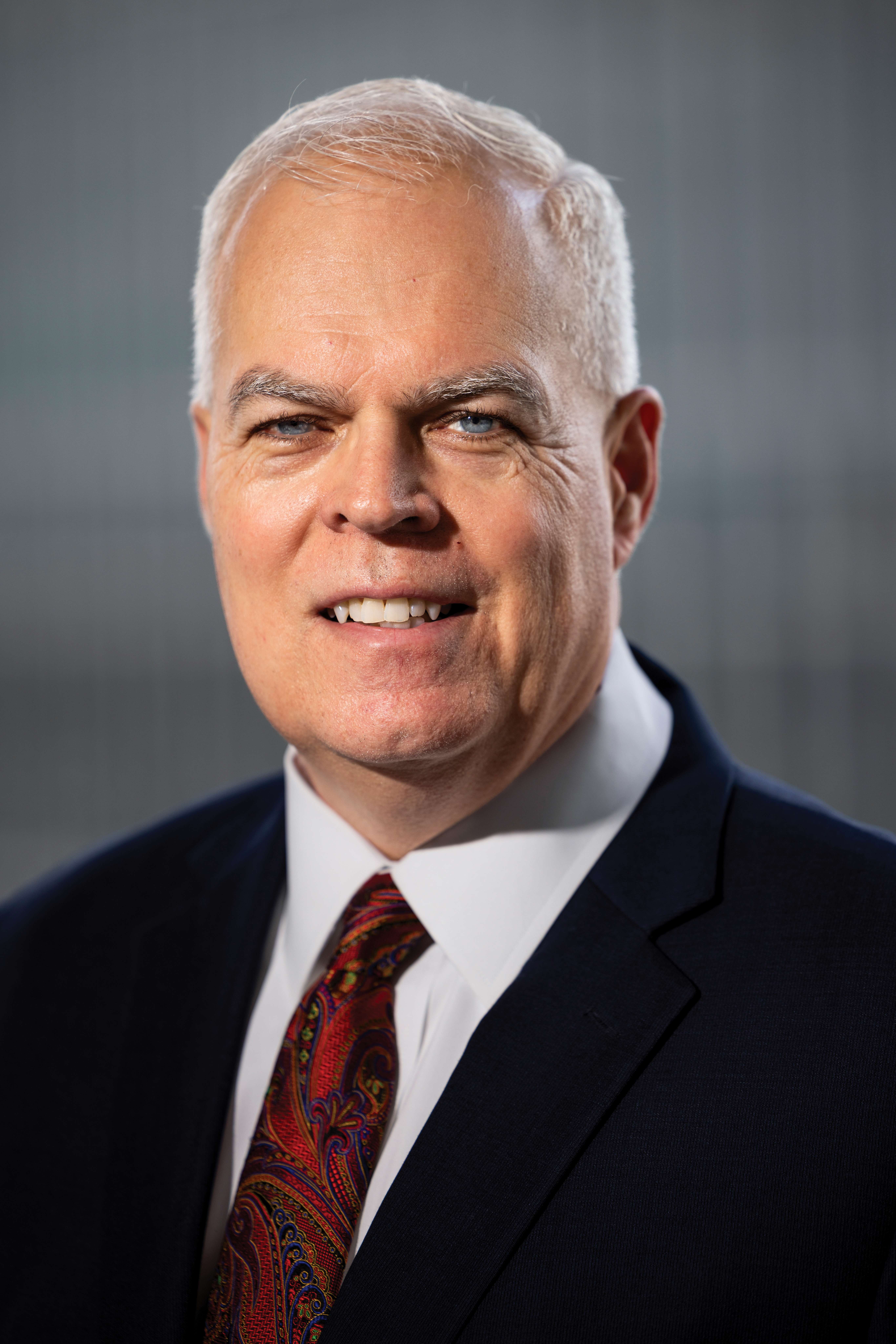 Professional headshot of a man in dark suit and red patterned tie smiling at camera