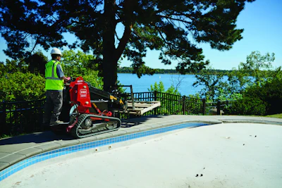 Worker in safety vest operating red stump grinder equipment beside residential pool with lake and trees in background