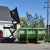 Worker operating green dumpster with red compactor truck in residential driveway on sunny day