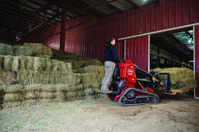 Red tracked mini loader positioned in barn with stacked hay bales and farm equipment