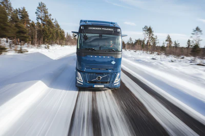 Blue Volvo hydrogen-powered truck driving on snowy road through forested landscape