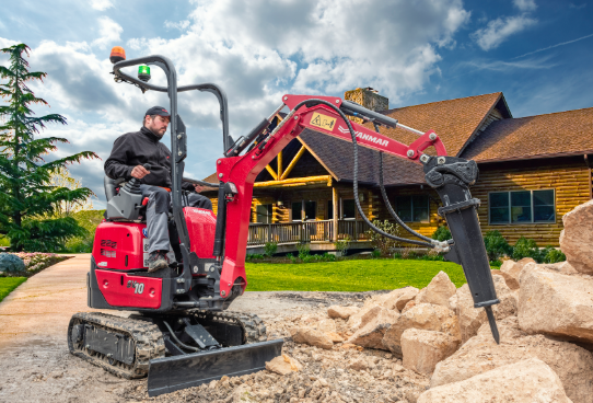 Red mini excavator with hydraulic arm operating on gravel near a rustic lodge building under blue sky