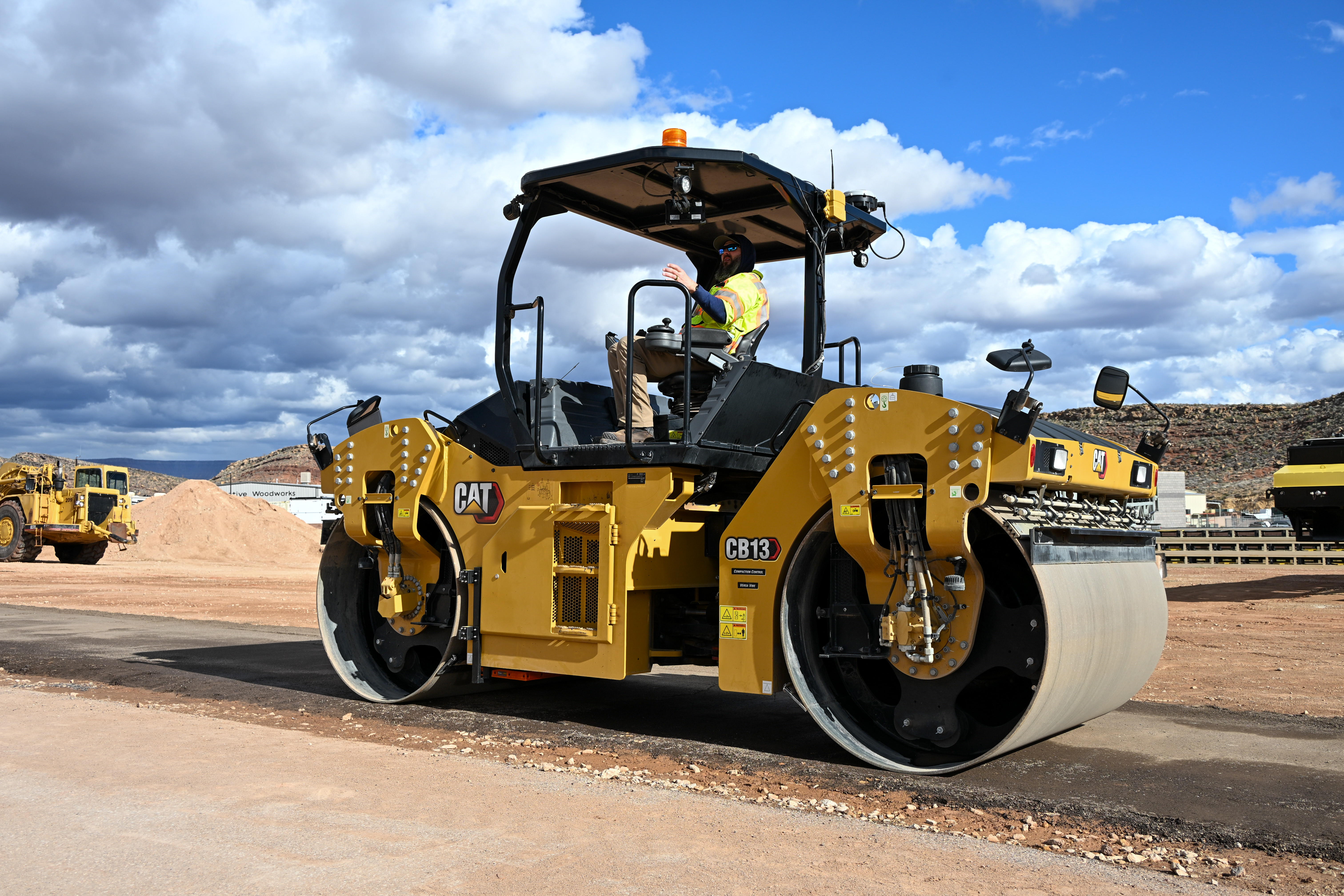 Yellow CAT CB13 asphalt roller compactor with operator on construction site under blue sky