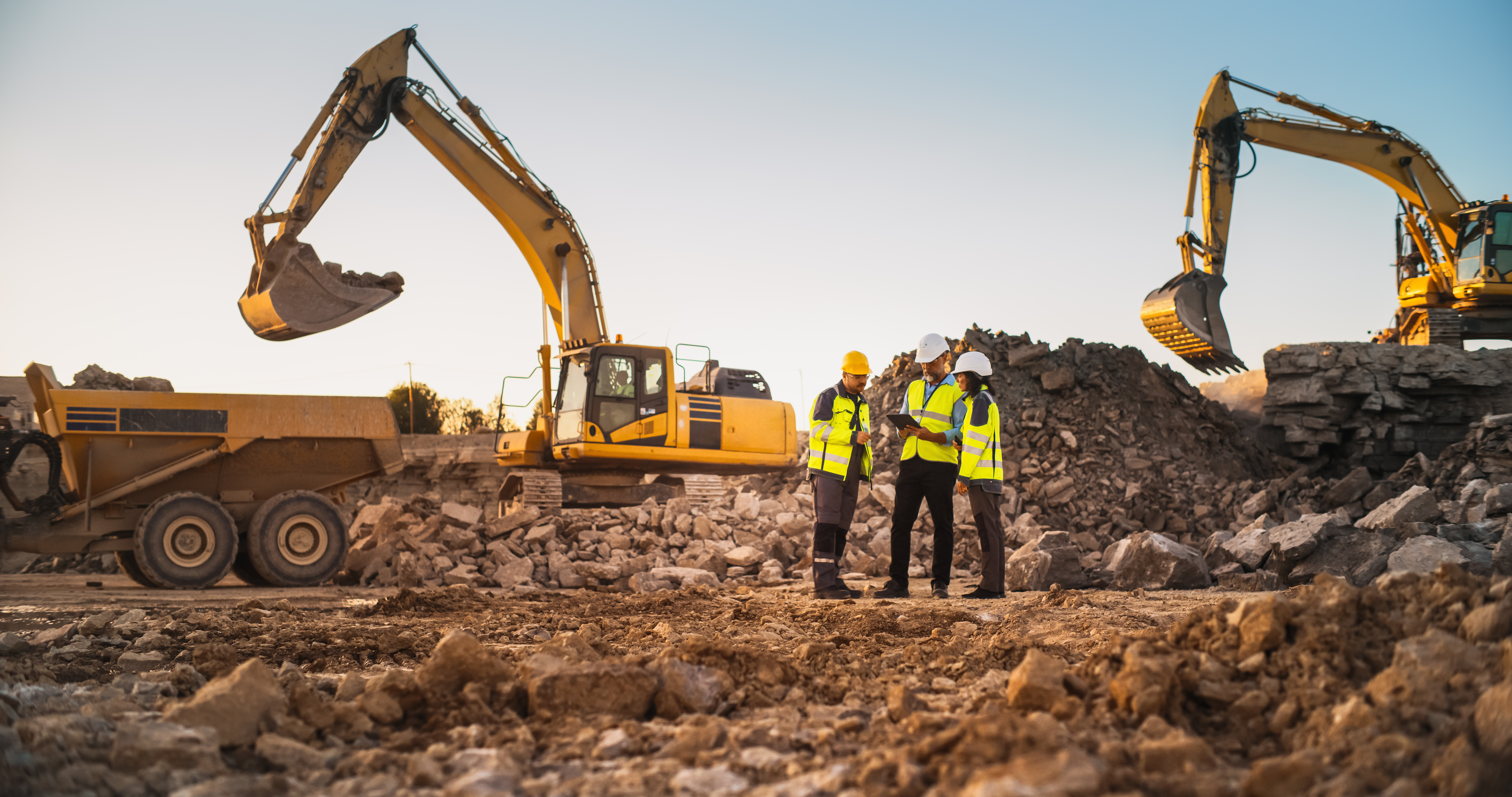 Construction site with two excavators, workers in safety vests, and dump trucks moving earth and rock materials