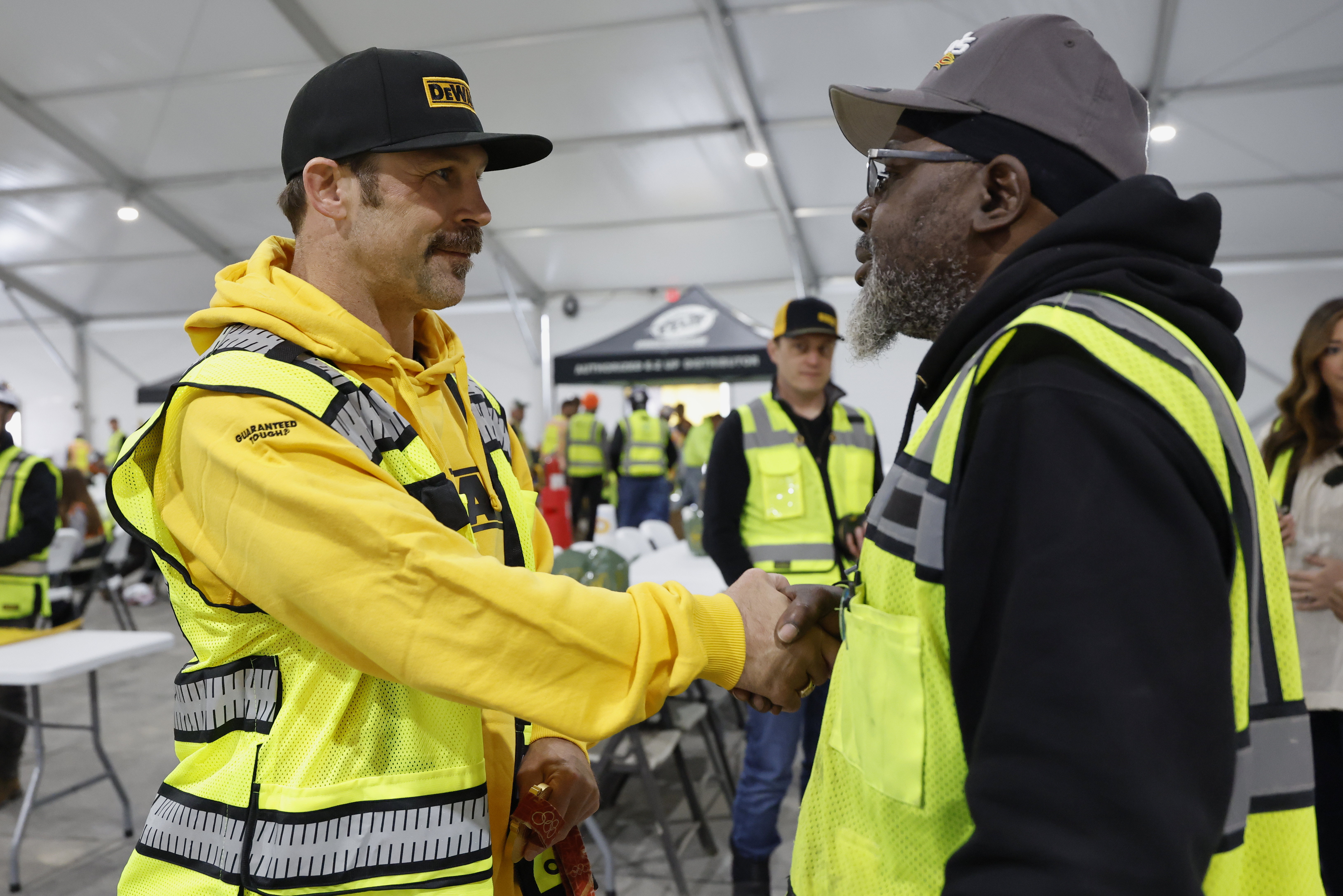 Two construction workers in safety gear shake hands during a workplace meeting in an industrial tent facility.