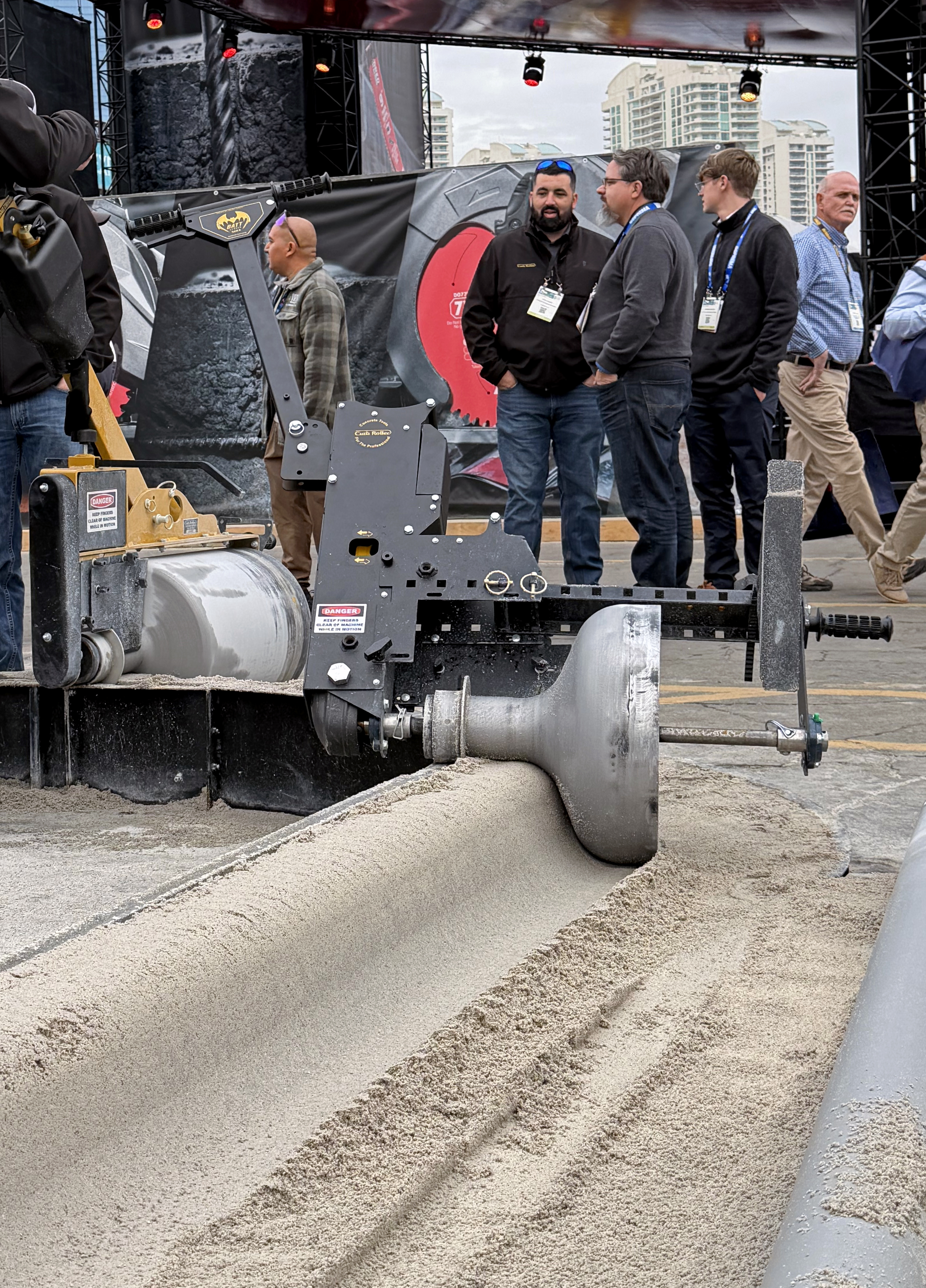 Industrial soil compactor machine displayed at outdoor trade show with attendees in hard hats observing the equipment