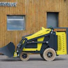 Yellow and black ECOVOLVE compact loader with bucket attachment positioned against wooden building
