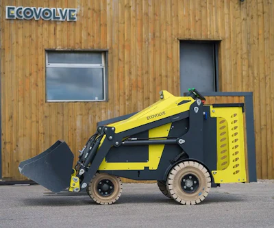 Yellow and black ECOVOLVE compact loader with bucket attachment positioned against wooden building