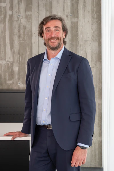 Professional headshot of a smiling man in navy blazer and light blue shirt against concrete wall backdrop