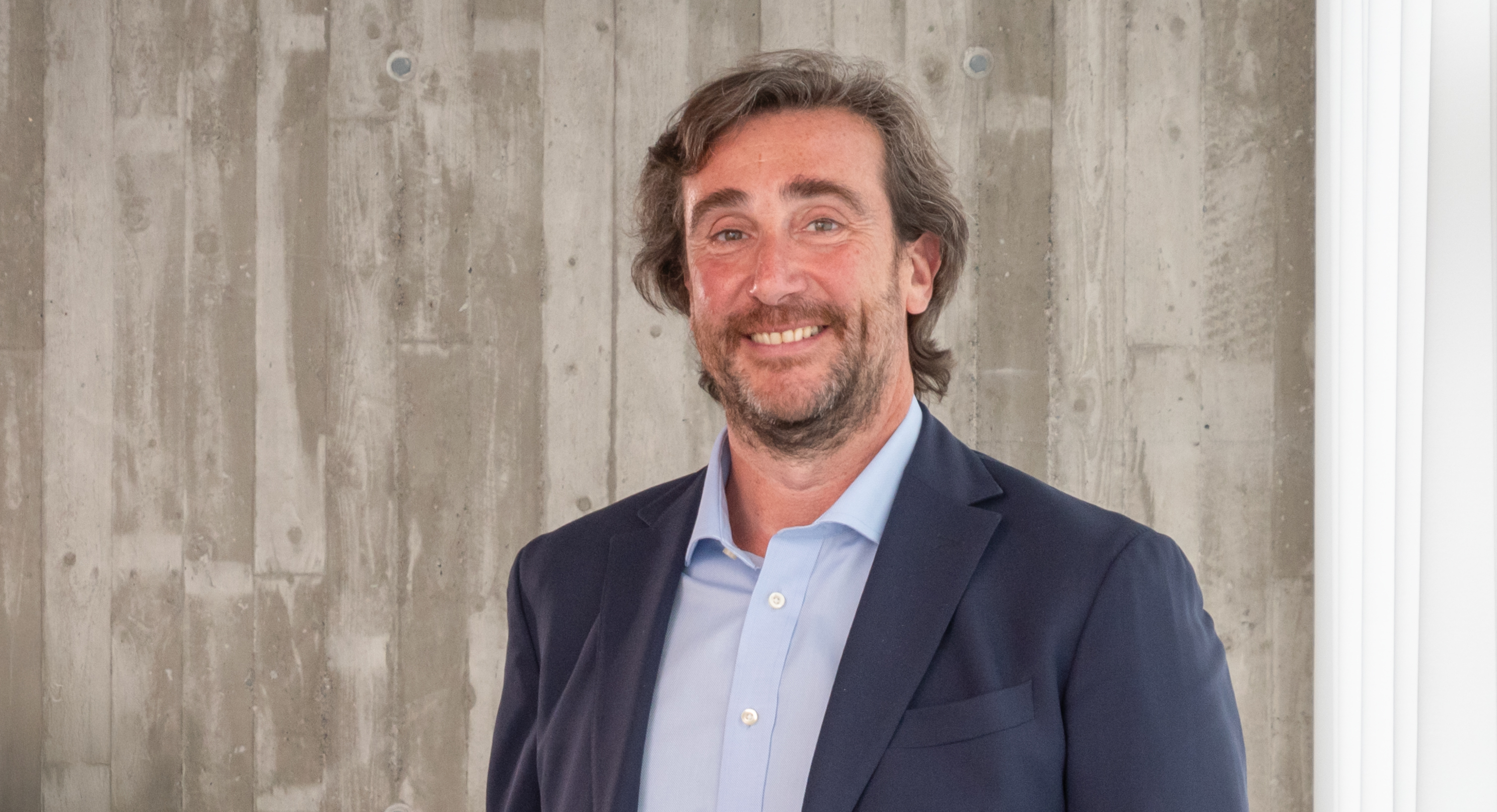 Professional man in navy blazer and light blue shirt smiling at camera against textured wall background