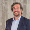 Professional man in navy blazer and light blue shirt smiling at camera against textured wall background
