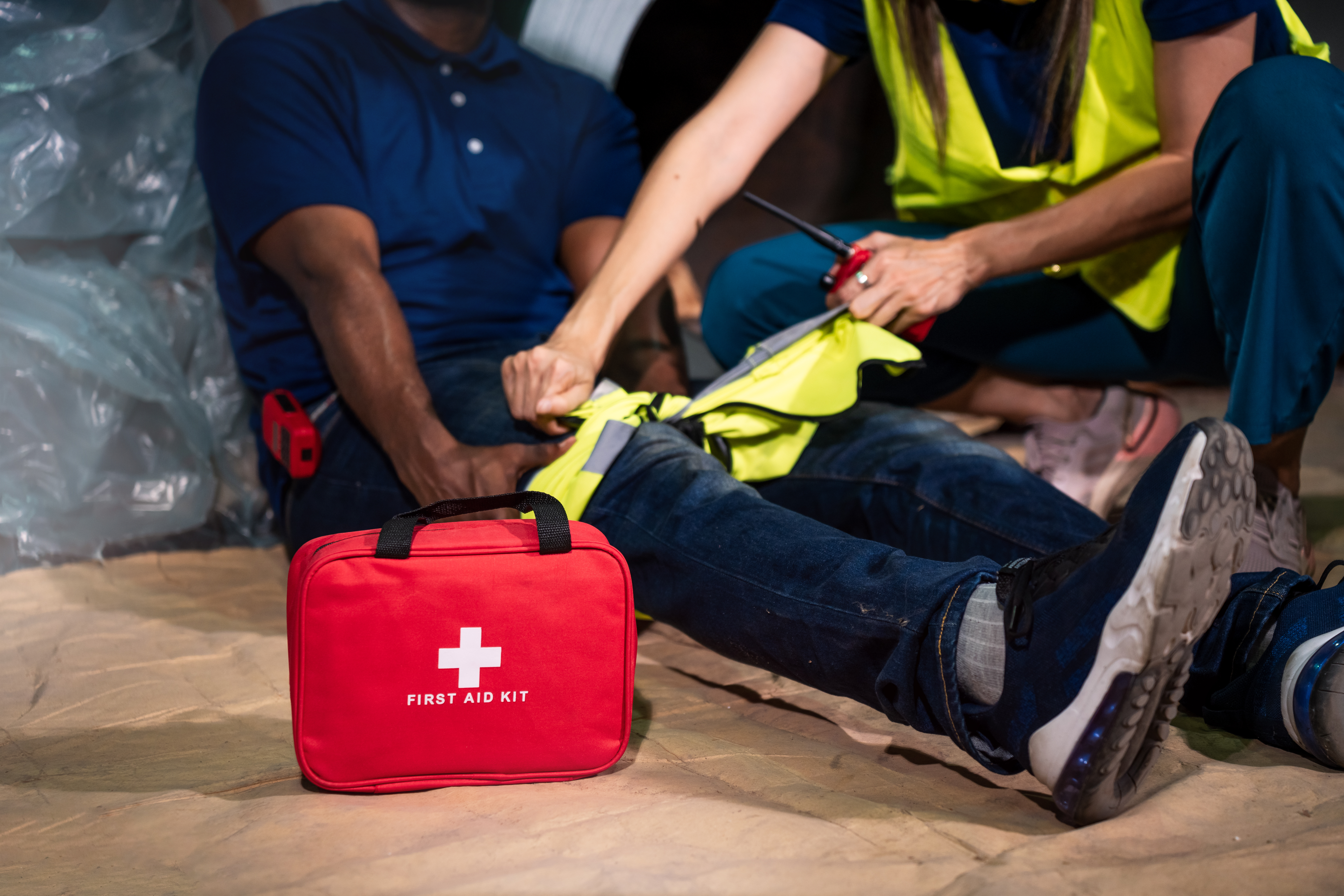 Workers in safety vests attending to an injured person with a red first aid kit nearby on the ground