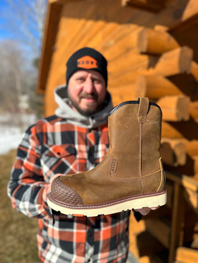 Man in plaid shirt holding tan Ariat work boot with cream-colored sole outdoors
