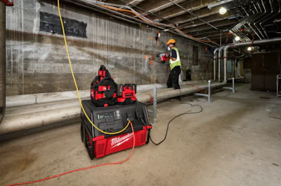Construction workers using Milwaukee power equipment in an industrial building interior with concrete walls and exposed ceiling infrastructure.