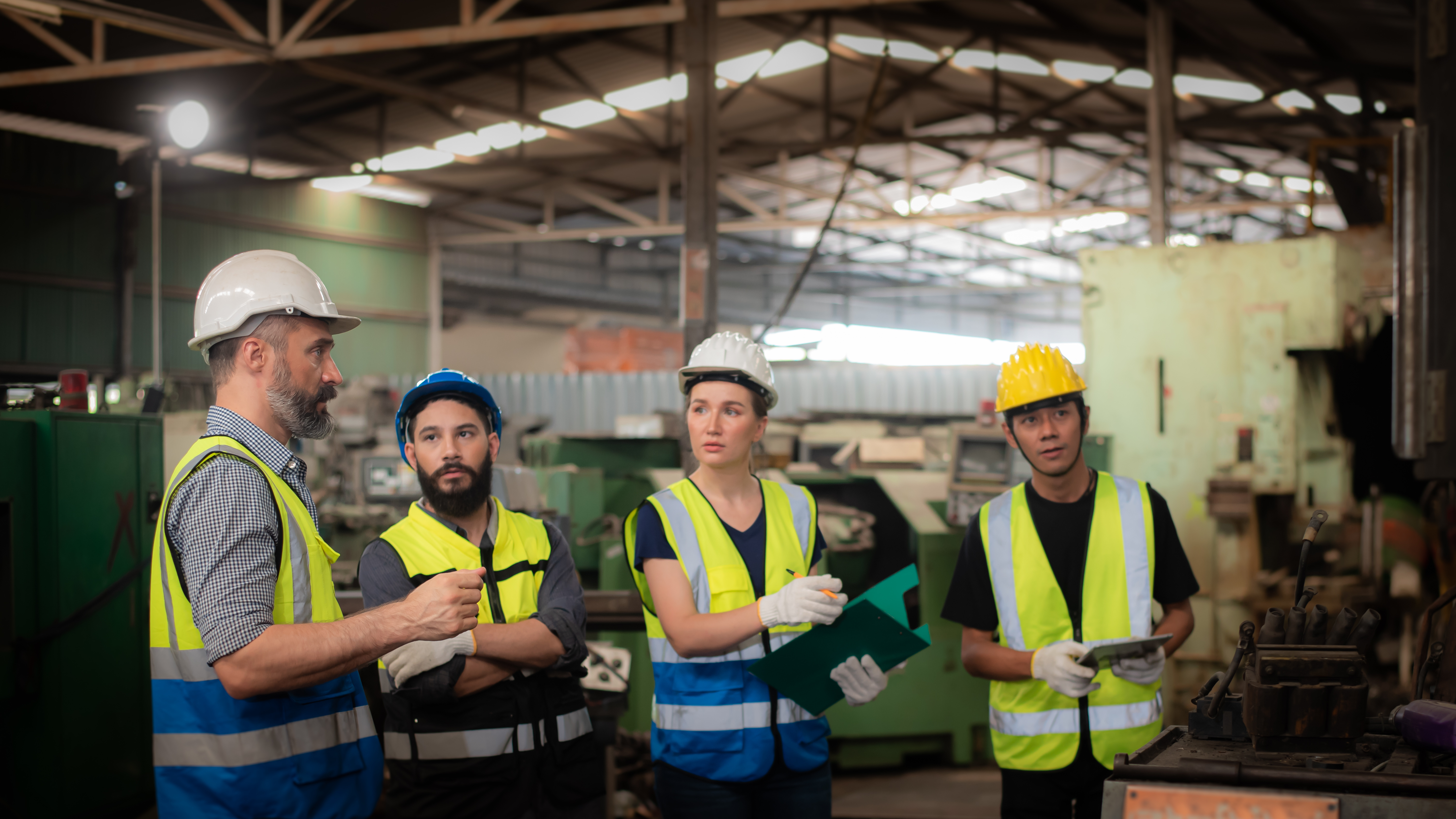 Four construction workers wearing safety vests and hard hats in an industrial warehouse discussing project plans