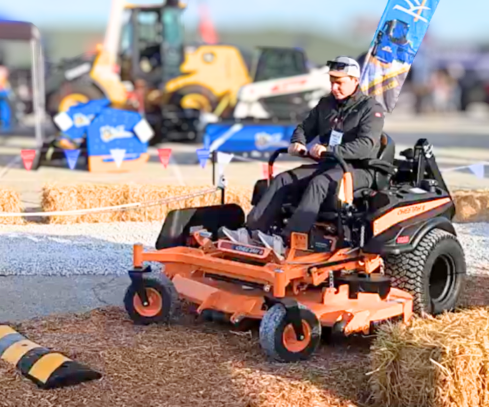 Operator demonstrates zero-turn mower at outdoor equipment event with construction machinery in background