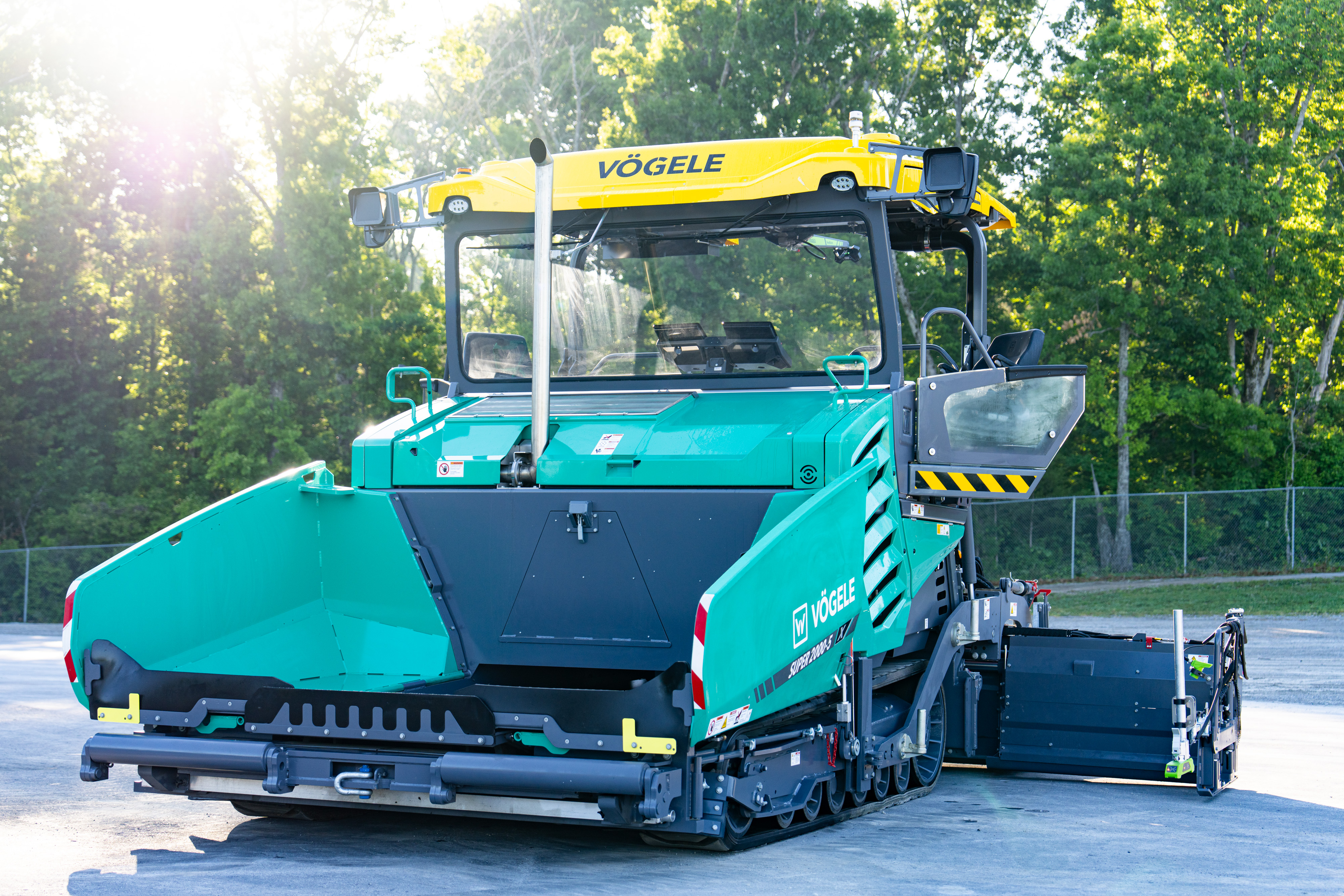 Turquoise VÖGELE asphalt paver with yellow crane attachment parked outdoors near trees