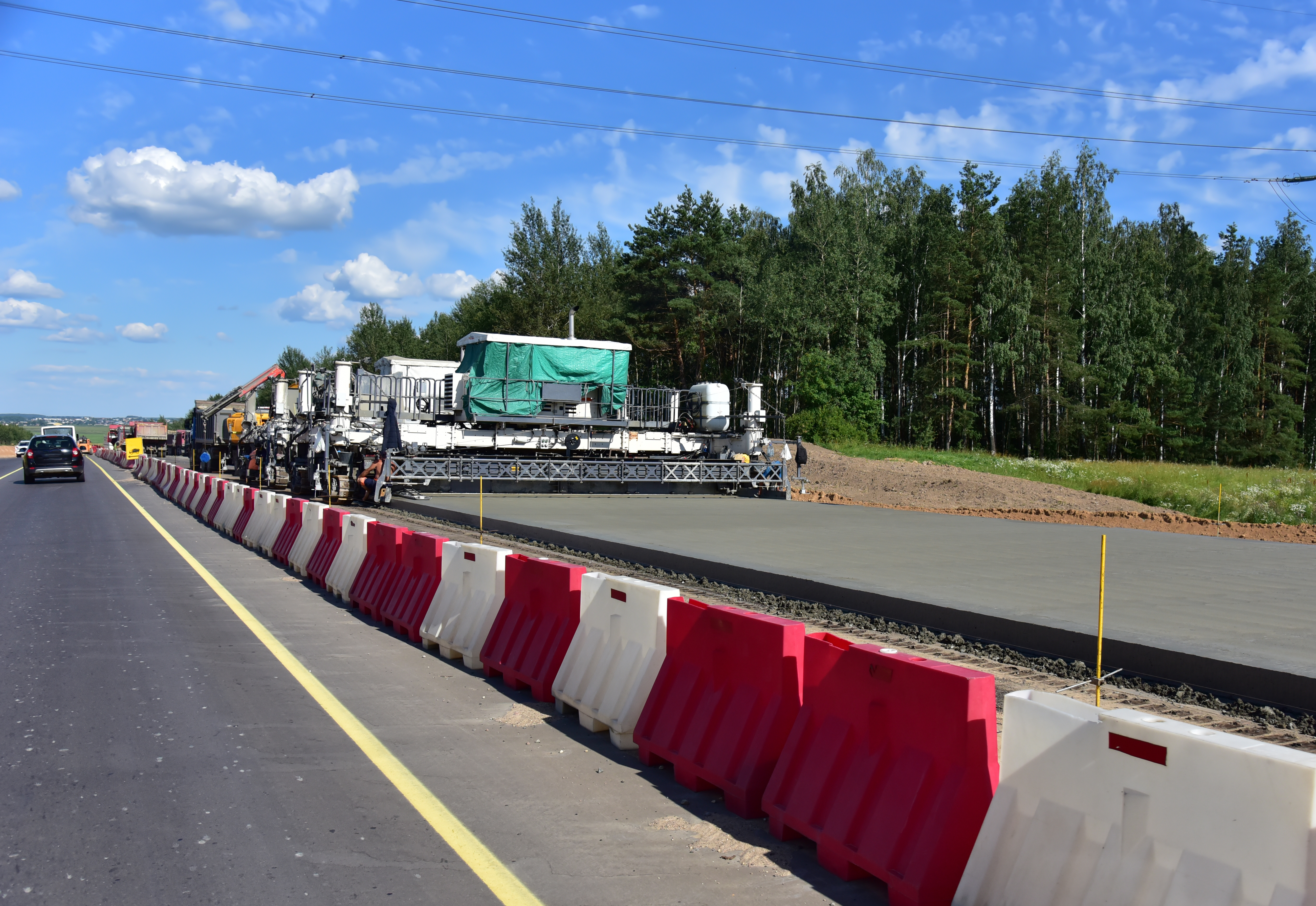 Highway construction site with concrete paving machine and red-white safety barriers along roadside