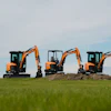 Three orange and black compact excavators lined up in a green field under a partly cloudy sky