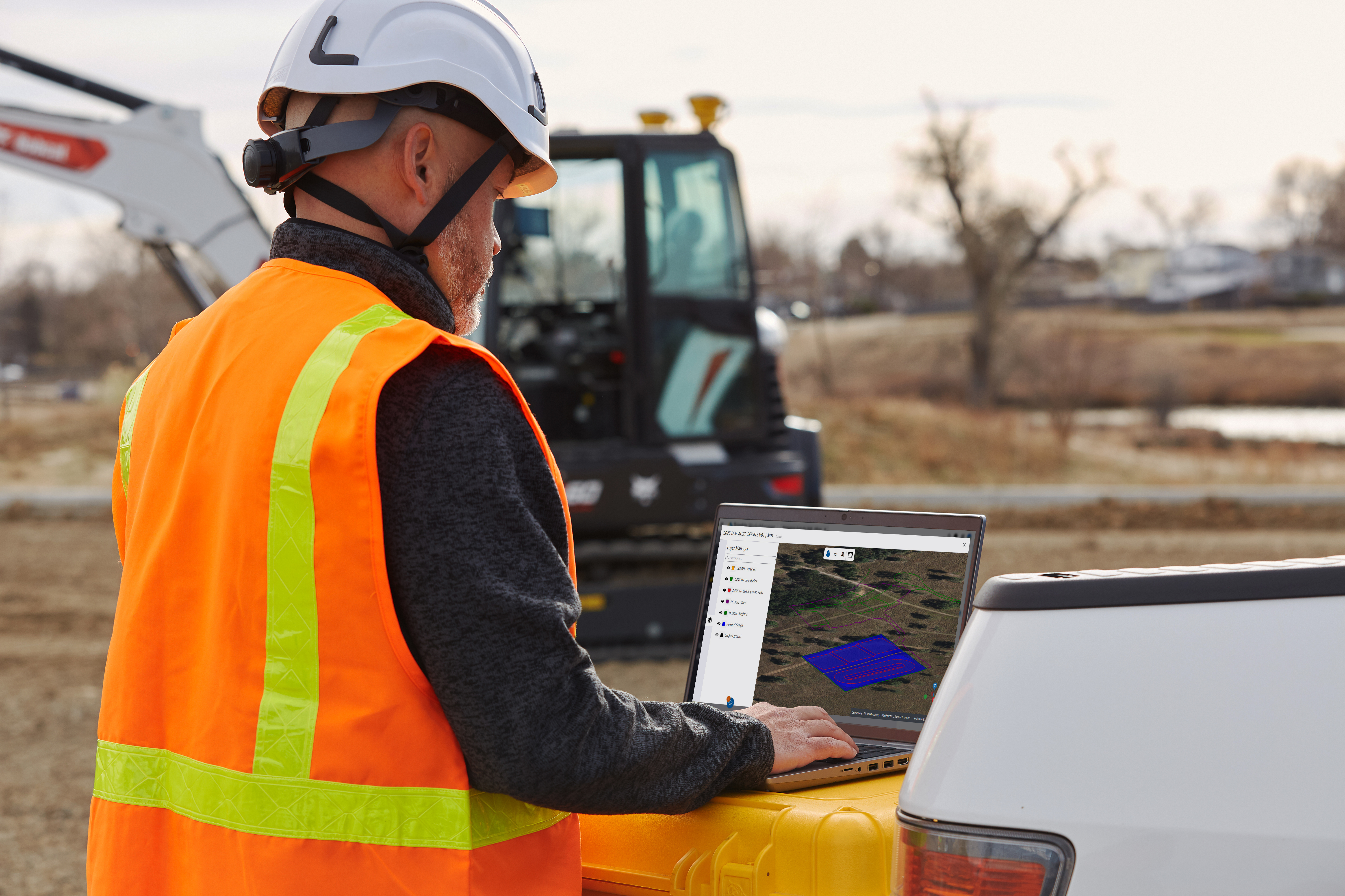 Construction worker wearing safety helmet and orange vest using laptop at outdoor job site with excavator in background