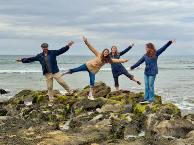 Five people with arms raised in celebration standing on moss-covered rocks at a beach with ocean waves in the background