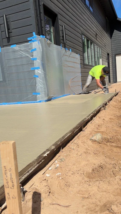 Construction worker in yellow safety vest pouring concrete foundation next to residential building