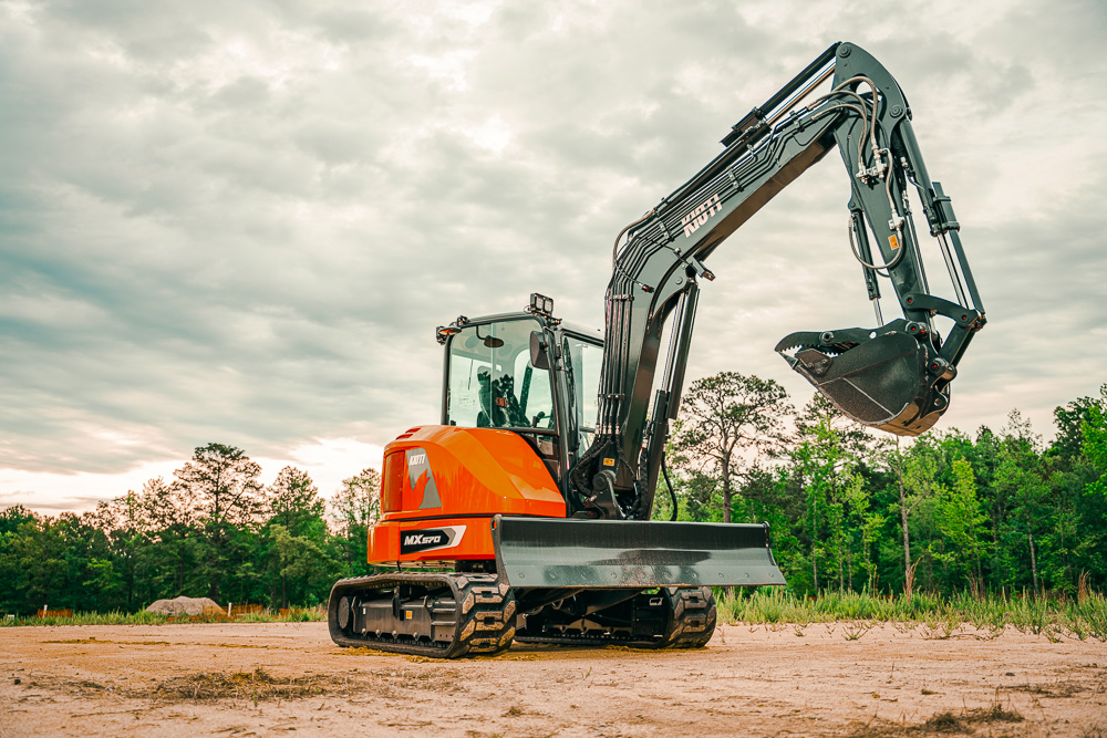 Orange Kubota mini excavator with extended boom on sandy construction site surrounded by trees