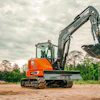 Orange Kubota mini excavator with extended boom on sandy construction site surrounded by trees